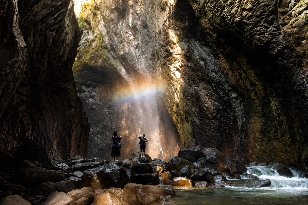 A waterfall falling into a narrow rock canyon between high cliff walls, a rainbow in the spray and two people standing on the rocks below.