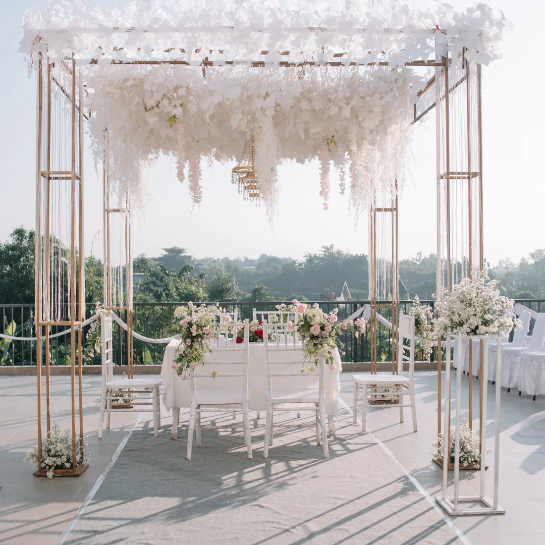 A rooftop pelaminan at Achiera under a canopy of hanging white florals, the couple’s table set beneath it and rows of white chairs over the treeline.